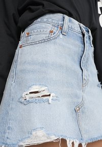 Jupe mini en denim bleu clair, avec détails déchirés et ourlet effiloché. Design classique à cinq poches avec des accents de quincaillerie en bronze.