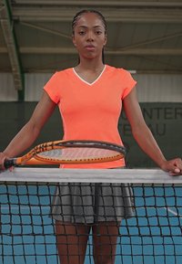Camisola atlética laranja brilhante com acabamento branco, acompanhada de uma saia cinza. Raquete de ténis segurada sobre uma rede em um ambiente de court coberto.