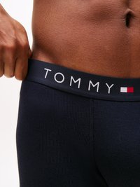 Mornarska elastična pasnica z napisom "TOMMY" v beli barvi; spodaj prilegajoča se ribana tekstura tkanine. Povečava srednjega dela, ki prikazuje gladko kožo.