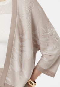 Beżowy sweter kardigan z dzianiny w paski, z szerokimi rękawami i kontrastowym, jednolitym wykończeniem. Teksturowana tkanina z subtelnymi detalami wzorniczymi.