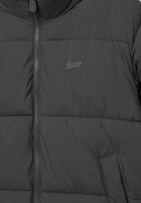 Sort puffjakke med lynlås, quiltet tekstur, elastisk hætte og en sidelomme med diskret logodetalje på brystet.