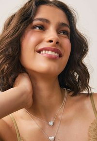 Colliers en forme de cœur en argent empilés, l'un brillant avec des strass et l'autre lisse. Le modèle a des cheveux bruns ondulés et porte un haut vert clair.