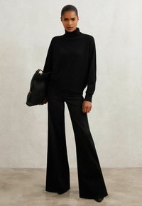 Musta poolne hakkide särk koos kõrge vöökohaga, laienevate mustade pükstega. Mudel hoiab suurt musta käsipakki, millel on kuldsed detailid, neutraalse tausta ees.