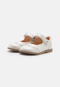 White patent leather shoes with a rounded toe, cut-out pattern, and a silver adjustable strap. Light brown sole, smooth texture.