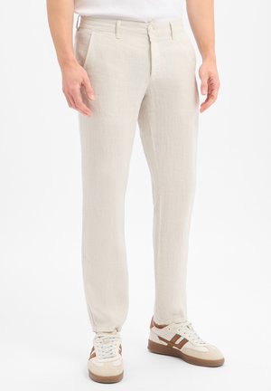 Man draagt lichtbeige broek met voorzakken en witte sneakers met bruine accenten, staand op een witte achtergrond.