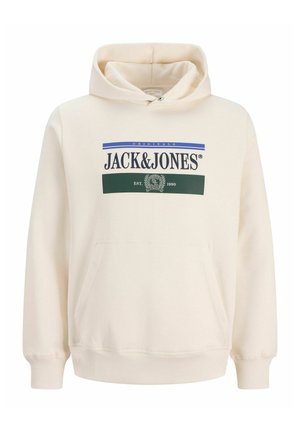 Ragazzo con capelli ricci che indossa una felpa crema Jack & Jones, camicia bianca, jeans blu a gamba larga e scarpe beige, in piedi davanti a uno sfondo bianco.