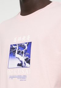 Gaiši rozā kokvilnas T-krekls ar Hokusai "Cīruļi uz sniegota koka" druku zilā krāsā. Baltais teksts zem grafikas apkopo mākslas darba detaļas.