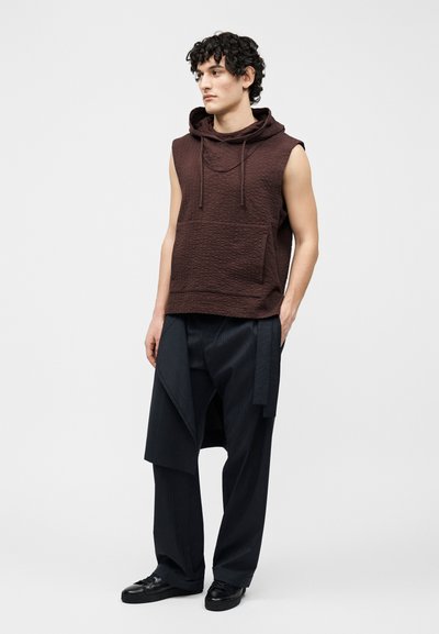 Jonge man met krullend haar, gekleed in een mouwloos bruin capuchonshirt en zwarte broek met ruime pijpen en stoflaag, staand met handen in de zakken.