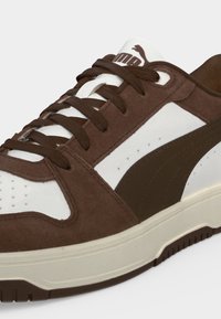 Καφέ και λευκό αθλητικό παπούτσι Puma με καφέ κορδόνια, καφέ σουέντ λεπτομέρειες και λευκή σόλα σε ανοιχτό φόντο.