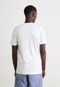 Weißes Baumwoll-T-Shirt mit rundem Halsausschnitt, kurzen Ärmeln und glatter Textur. Trägt eine Person, die von hinten gesehen wird, kombiniert mit blauen Cargo-Hosen.