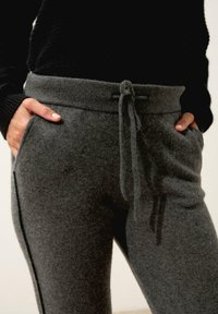 Šedé pletené tepláky s pasem na šňůrku, bočními kapsami a měkkým, texturovaným povrchem. Obsahují kontrastní černé boční pruhy.