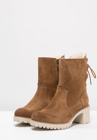 Bottines en daim marron avec des talons épais beige, fermeture éclair sur le côté, doublure en fourrure et détail en dentelle à l'arrière, sur un fond blanc uni.