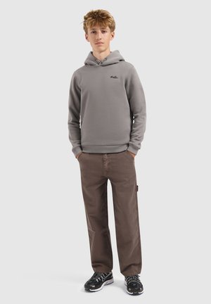 Tienerjongen staat met zijn handen in zijn zakken, draagt een grijze hooded sweatshirt, bruine broek en zwarte sneakers op een effen achtergrond.