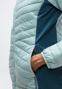 Hånd i sidelommen på lys blå quiltet jakke med mørk blå stoffpanel og glidelåsdetalj.