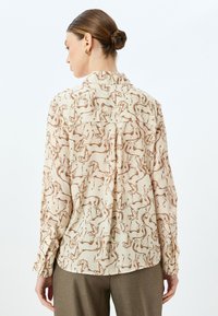 Lange mouw blouse van lichtbeige stof met een bruine paardenprint. Bevat een kraag en knoopmanchetten, met een iets losse pasvorm.