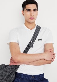 T-shirt blanc à manches courtes avec le logo Lee, associé à un pantalon bleu foncé. Sac à bandoulière à carreaux gris porté sur l'épaule, doté d'une boucle en métal.