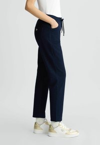 Pantalons de jogging bleu marine avec taille élastique, cordon de serrage, poches latérales et texture mouchetée. Associés à des baskets blanches avec des accents dorés.