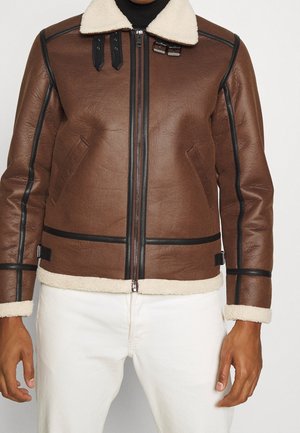 Veste en cuir marron avec bordure noire et col, poignets et ourlet en peau lainée blanche, portée avec un pantalon blanc.