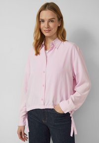 Kobieta o blond włosach do ramion, ubrana w jasnoróżową koszulę zapinaną na guziki i ciemne dżinsy, stojąca na jednolitym szarym tle.