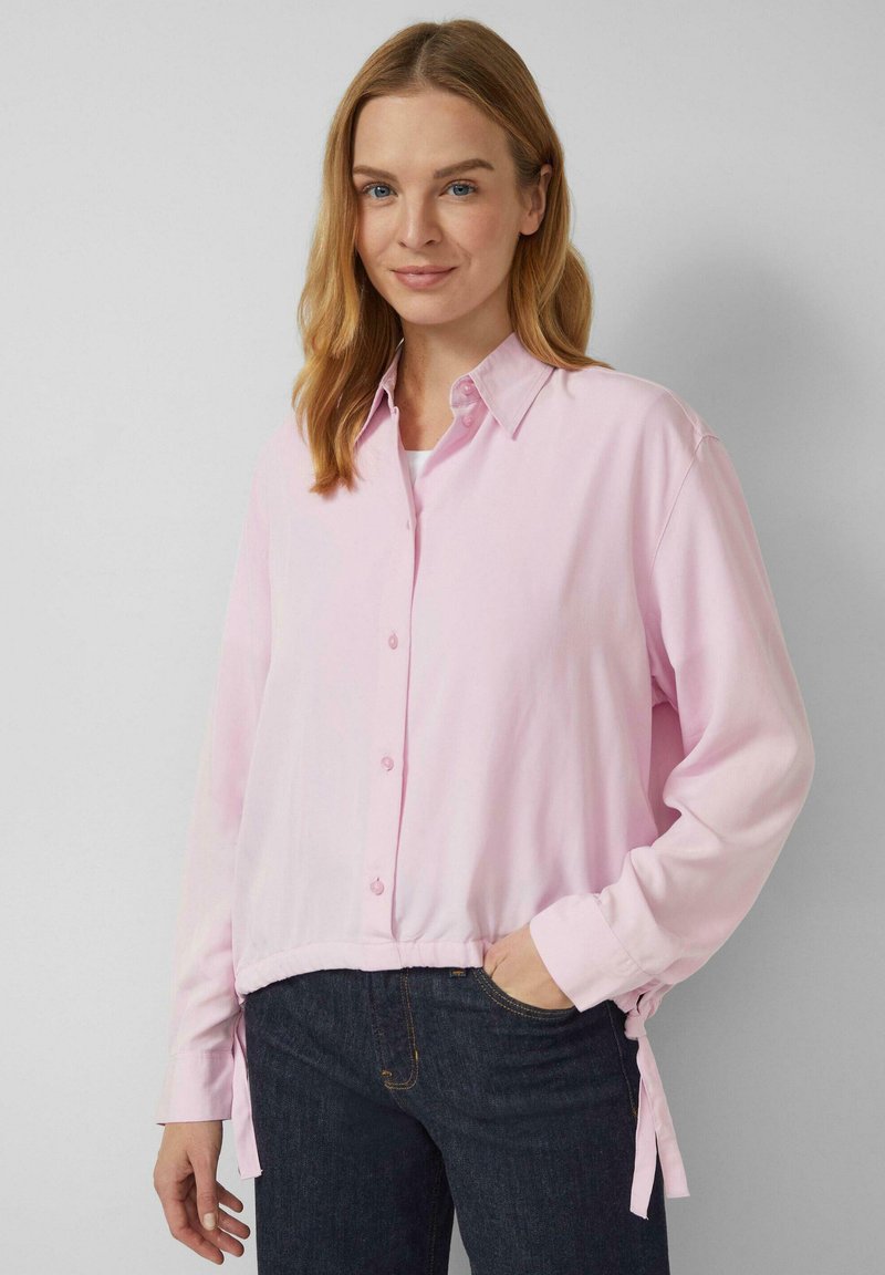 Kobieta o blond włosach do ramion, ubrana w jasnoróżową koszulę zapinaną na guziki i ciemne dżinsy, stojąca na jednolitym szarym tle.