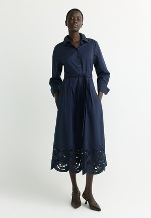 Woman wearing a long-sleeve navy blue shirt dress with a tied waist and decorative cutouts at the hem, standing with hands in pockets.