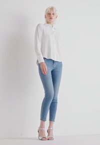 Camicia bianca a maniche lunghe con bottoni, abbinata a jeans skinny blu chiaro e sandali con tacco argento, su uno sfondo bianco semplice.
