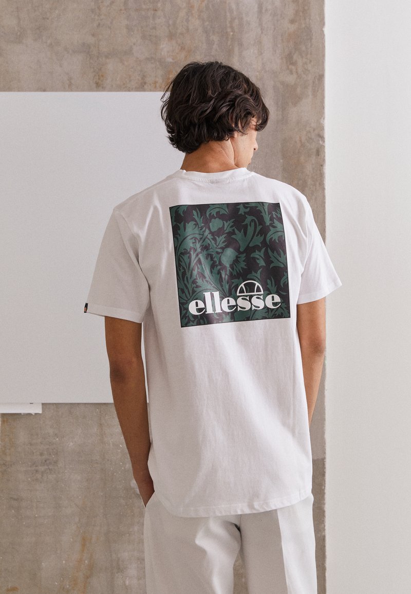Valge puuvillane t-särk, mille seljal on ristkülikukujuline must ja roheline lilleline graafika, mille all on tekst "ellesse". Lühikesed varrukad.