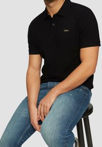 Polo noir avec un logo doré discret sur la poitrine, associé à un jean en denim bleu clair, mettant en valeur une coupe classique et une texture lisse.