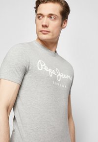 Pelēks īsām piedurknēm T-krekls, izgatavots no mīksta kokvilnas, ar baltu apdrukātu logo "Pepe Jeans London" uz priekšpuses.
