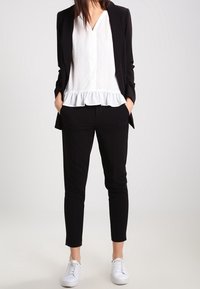 Schwarze Blazer mit einer weißen Bluse mit gerafftem Saum, kombiniert mit schwarzen Hosen und weißen Sneakern. Glattes Material, taillierte Passform.