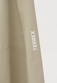 Μπεζ ύφασμα με κατακόρυφο λευκό λογότυπο "TERREX" τυπωμένο στη δεξιά πλευρά, που εμφανίζει υφασμάτινες λεπτομέρειες ραφών.
