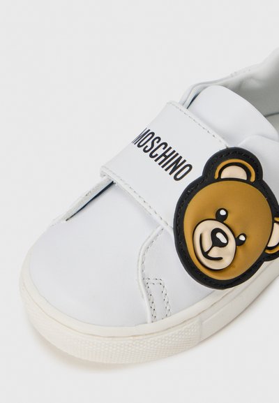Sneaker en cuir blanc présentant une application en forme de visage d'ours brun rond, sangle réglable avec le logo "Moschino" et semelle en caoutchouc texturée.