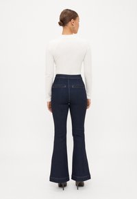 Calças flare de denim azul escuro com uma cintura alta, apresentando bolsos traseiros e costura em contraste, emparelhadas com uma blusa ajustada de mangas longas brancas.