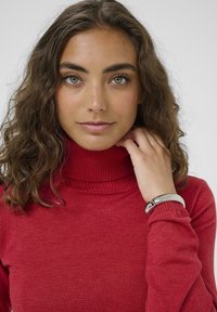 Piros garbónyakú pulóver ribbed textúrával és mandzsettával, ezüst színű karkötővel viselve. A modell hullámos, vállig érő hajjal rendelkezik.