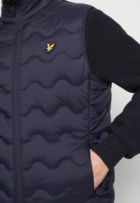 Mörkblå puffärmad väst med vågquiltmönster, dragkedjestängning och stickade ärmar. Har en liten gul fågel-logotyp på bröstet.