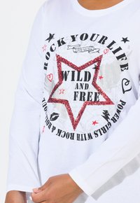 Valkoinen pitkähihaisen t-paita, jossa on punainen glitteritähti ja teksti "WILD AND FREE", sekä erilaisia mustia ja punaisia painatuksia.