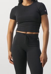 Kobieta ubrana w dopasowany czarny krótki T-shirt i czarne legginsy z wysokim stanem i sznurkiem w pasie, stojąca na jednolitym tle.