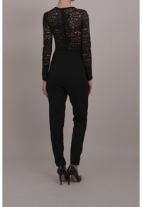 Czarny kombinezon z długimi koronkowymi rękawami, dopasowanym biustem i zwężającymi się spodniami. Teksturowane detale koronkowe na rękawach, połączone z wysokimi obcasami.
