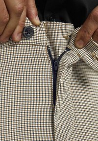 Vävda byxor har ett mönster i tan och brunt houndstooth, med dragkedja fram och svarta knappdetaljer vid midjan.