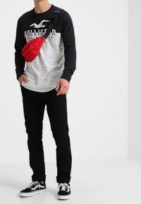 Pitkähihainen musta ja harmaa paita, jossa on "Hollister"-logo, mustat housut ja musta-valkoiset tennarit; punainen olkalaukku olalla.