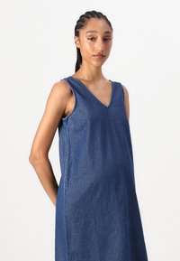 Vestido de ganga em azul escuro com decote em V, design sem mangas e um corte descontraído. O tecido tem um ligeiro brilho com costuras visíveis.