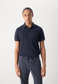 Polo shirt marine avec un col classique, des manches courtes et un petit logo. Associe à un pantalon à taille élastique gris foncé. Texture lisse.