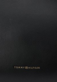 Černý texturovaný kožený povrch s malým zlatým logem Tommy Hilfiger umístěným ve středu blízko spodního okraje.