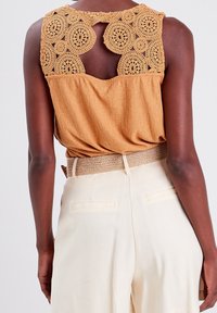 Top sans manches de couleur rouille avec des détails en dentelle délicate au dos. Associé à un pantalon crème avec une ceinture tissée et des poches latérales.