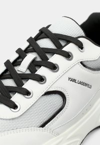 Λευκά αθλητικά παπούτσια με γκρι πάνελ από δίχτυ, μαύρα κορδόνια και μεταλλικά περβάζια. Διαθέτουν υφή στην επιφάνεια και ένα έντονο λογότυπο στο πλευρό.