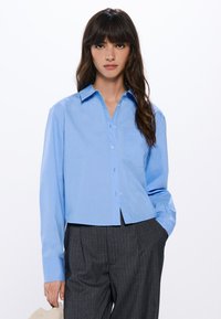 Camicia blu chiaro con maniche lunghe, colletto e taschino sul petto. Il tessuto è liscio e leggermente strutturato, con lunghezza che termina sopra la vita.