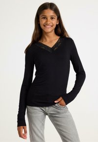 Sort langærmet skjorte med V-hals og blondekant. Blødt stof med en tætsiddende pasform, parret med lysegrå jeans.