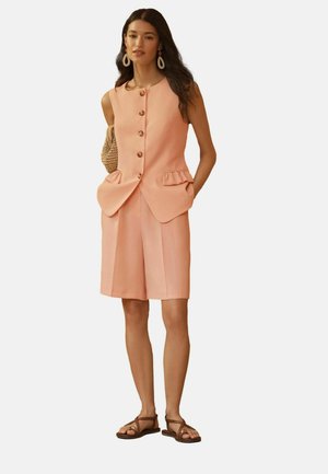 Frau trägt eine ärmellose, pfirsichfarbene Weste mit Knöpfen und Rüschen an den Taschen, passende knielange Shorts, braune Sandalen und eine geflochtene Handtasche.