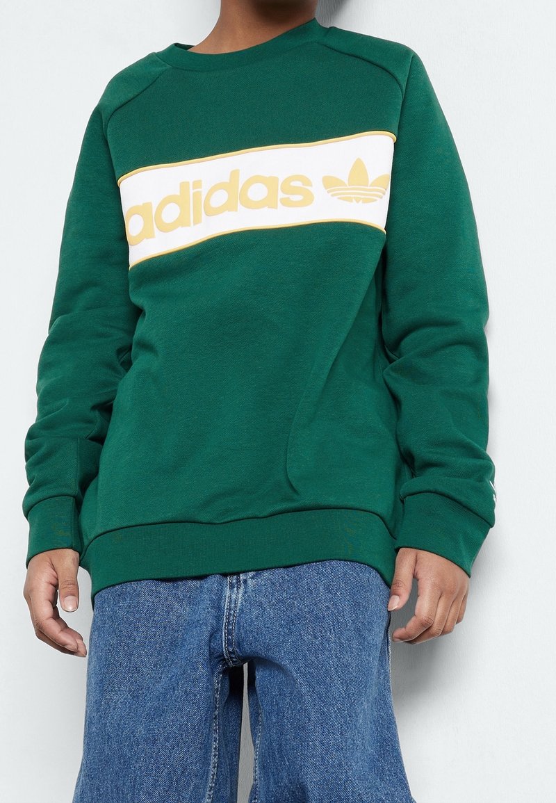 Grön sweatshirt med ett vitt horisontellt rand med "adidas" i gult och logotyp. Ribbad mudd och nederkant med en texturerad ytfinish.