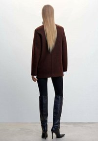 Pull marron oversize à coupe droite, porté avec des leggings noirs et des bottes noire à talons aiguilles mi-mollet, vu de dos.